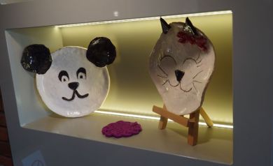 Na zdjęciu w oświetlonej wnęce dekoracyjne elementy ceramiczne przypominające kota i psa. Po środku leży również serwetka w kolorze fuksji.  - powiększ