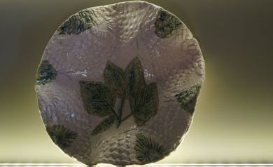 Na zdjęciu element ceramiczny, o nieregularnych kształtach, zdobiony zielonymi wzorami liści i nierówna fakturą.  - powiększ