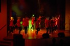 Na zdjęciu widać grupę dzieci lub młodych osób, które tańczą na scenie. Są ubrani prawdopodobnie w czerwone koszulki i ciemne spodnie, mają również jasnoniebieskie opaski na głowach. Scena jest oświetlona reflektorami i kolorowym światłem.