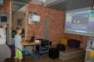Trzej chłopcy grający w grę komputerową wyświetlaną na projektorze. Wokół widać sprzęt elektroniczny i ceglaste ściany. 