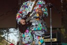 Mężczyzna w kolorowym garniturze znajduje się na scenie i gra na gitarze. Wokół niego sprzęt muzyczny. 