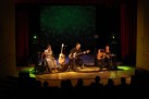 Na zdjęciu widać koncert zespołu Renata Przemyk TRIO, odbywający się na scenie w sali widowiskowej. Muzycy grają na gitarach i innych instrumentach i śpiewają przy nastrojowym oświetleniu. Widać również część publiczności pogrążonej w ciemności.
