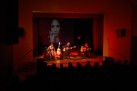 Na zdjęciu widać koncert zespołu Renata Przemyk TRIO, odbywający się na scenie w sali widowiskowej. Muzycy grają na gitarach i śpiewają przy nastrojowym oświetleniu. Na ekranie za muzykami wyświetlane jest zdjęcie kobiety. Widać również część publiczności.