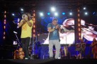 Na pierwszym planie dwaj muzycy, jeden grana trąbce, drugi na puzonie. W tle inni muzycy wraz z instrumentami. Całość ma miejsce na scenie plenerowej. 