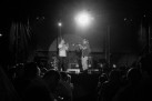 Czarno-białe zdjęcie muzyków występujących na scenie (jeden gra na trąbce, drugi śpiewa) oraz części publiczności. 