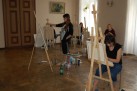 Na zdjęciu widać grupę osób, prawdopodobnie uczestników warsztatów plastycznych, malujących przy sztalugach w przestronnym pomieszczeniu. W pokoju z drewnianą podłogą i dużymi oknami znajduje się kilka sztalug z płótnami. Kobiety w różnym wieku malują, używając farb i pędzli. Na ścianie w tle wisi obraz. 