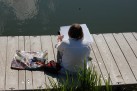 Na zdjęciu widać osobę siedzącą na drewnianym pomoście nad wodą, która zajmuje się malowaniem lub rysowaniem. Obok niej leży torba z przyborami artystycznymi. 
