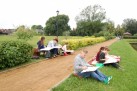 Na zdjęciu widać grupę młodych ludzi, biorących udział w plenerze malarskim lub rysunkowym w parku. Część osób z zdjęcia siedzi na ławce, podczas gdy inni rozłożyli się na kocach na trawie, tuż obok żwirowej ścieżki. Wszyscy mają przed sobą białe bloki do rysowania lub podkłady malarskie. Tło stanowi zielony park z zadbanym trawnikiem, krzewami i drzewami. W oddali widać budynki, a wzdłuż ścieżki rosną ozdobne krzewy. Po prawej stronie zdjęcia znajduje się niewielki zbiornik wodny.
