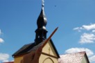 Na zdjęciu widać rękę trzymającą ołówek tak, że jego czubek wydaje się dotykać wieży kościoła w tle. Budynek stoi pod błękitnym, słonecznym niebem z kilkoma chmurami.