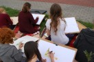 Zdjęcie przedstawia grupę ludzi, uczestników warsztatów, biorących udział w plenerze malarskim. Malują lub rysują na zewnątrz, siedząc na kocach lub matach na trawie, z podkładkami i przyborami malarskimi. Wszyscy są pochyleni nad swoimi pracami, które wydają się przedstawiać budynki.
