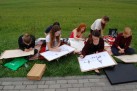Na zdjęciu widać grupę osób, siedzących na trawie i rysujących. Osoby znajdują się na zewnątrz, na trawiastym terenie, obok ścieżki. Uczestnicy siedzą, trzymając podkładki do rysowania i szkicując. Wokół nich leżą przybory malarskie i inne przedmioty. W grupie jest kilka młodych osób, które są skupione na swoich pracach.