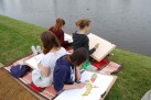 Zdjęcie przedstawia cztery młode osoby, siedzące na kocu na trawiastym brzegu zbiornika wodnego. Każda z nich ma przed sobą podkładkę do rysowania i zajmuje się szkicowaniem lub malowaniem.