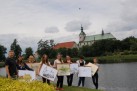 Zdjęcie przedstawia grupę osób, prezentujących swoje prace plastyczne na tle zbiornika wodnego, roślinności, kościoła i innych budynków. 