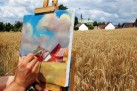 Na obrazku widać artystę malującego obraz w plenerze. Na pierwszym planie ręce trzymające pędzel i paletę, malujące na sztalugach. Na płótnie powstaje obraz przedstawiający wiejski pejzaż. Tło stanowi pole dojrzałego zboża, z pojedynczymi, czerwonymi makami. W oddali widać zabudowania, w tym domy z białymi fasadami i wysoką wieżę kościoła. Niebo jest pochmurne, z widocznymi, białymi chmurami.