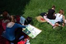 Na zdjęciu widać grupę młodych ludzi siedzących na trawie, na kocach - malują i rysują. Jedna z osób maluje obraz przedstawiający budynek.