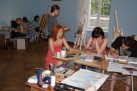 Na zdjęciu widać grupę osób uczestniczących w warsztatach plastycznych. Uczestnicy siedzą przy stołach lub pracują przy sztalugach w jasnej pracowni. Na stołach znajdują się różne przybory artystyczne, takie jak farby, pędzle, szkicowniki i inne materiały plastyczne. W tle widać inne osoby zajmujące się swoimi pracami.