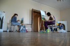 Na zdjęciu widać trzy osoby, uczestniczki warsztatów, malujące obrazy w dużej, jasnej sali. Każda z nich siedzi na stołku przed sztalugą i jest skupiona na swojej pracy. W pomieszczeniu znajdują się różne akcesoria malarskie, takie jak farby, pędzle i pojemniki. Podłoga jest pokryta wzorzystymi płytkami, a w tle widać otwarte drzwi prowadzące do innego pomieszczenia.