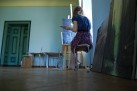 Na zdjęciu widać osobę siedzącą na stołku i malującą obraz na sztaludze. Scena rozgrywa się w jasnym pomieszczeniu, prawdopodobnie pracowni malarskiej, z błękitnymi ścianami. W tle widoczne są inne obrazy, w tym duży, ciemny pejzaż oparty o ścianę. Obok artysty stoi butelka wody, a w oddali widać stoliki z różnymi przedmiotami oraz zamknięte drzwi.