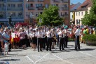 Orkiestra dęta w jednakowych strojach. Wokół inne osoby biorące udział w przemarszu, budynki i roślinność. W tle widać również pomnik. 