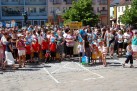 Tłum dorosłych i dzieci stojący na placu. Trzymają banery między innymi z nazwami szkół. W tle roślinność i zabudowa miejska. 