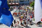 Zdjęcie zrobione z lotu ptaka. Na pierwszym planie powiewające flagi, na drugim maszerujący tłum ludzi, zaś w tle zabudowa miejska. 