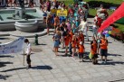 Na zdjęciu widać barwny pochód dzieci i dorosłych idących przez miejski plac, niosących transparenty i kolorowe dekoracje.