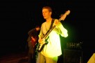 Mężczyzna grający na gitarze elektrycznej. Oświetla go żółte światło. W tle inny mężczyzna gra na gitarze basowej. 