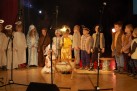 Na zdjęciu widać dzieci występujące w jasełkach, tradycyjnym przedstawieniu bożonarodzeniowym. Dzieci są na scenie w kostiumach, które symbolizują postacie związane z historią narodzin Jezusa, takie jak anioły, królowie i pasterze. 