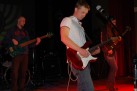 Na zdjęciu trzej mężczyźni grający na gitarach elektrycznych i na gitarze basowej. 