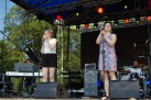 Dwie kobiety śpiewające do mikrofonów ustawionych na stojakach. Wokół nich sprzęt muzyczny, a także muzycy grający na keyboardzie i perkusji. Całość odbywa się na scenie plenerowej. 