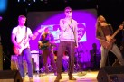 Na zdjęciu mężczyzna w ciemnych okularach śpiewa do mikrofonu, a także trzej mężczyźni grający na gitarach. 