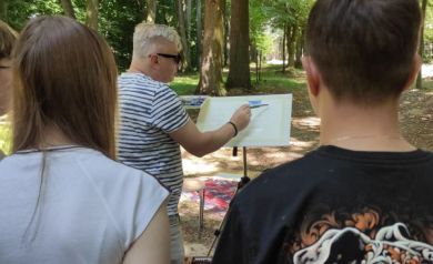 Na zdjęciu widoczne są dwie osoby stojące tyłem. Przed nimi mężczyzna w pasiastej koszulce i okularach przeciwsłonecznych maluje pędzlem obraz ustawiony na specjalnym stojaku. Wszystkie osoby z zdjęcia znajdują się w miejscu, gdzie jest dużo zieleni, m.in. są tam drzewa. 