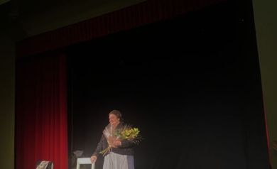 Aktorka na scenie z kwiatami. Opiera się o białe krzesło.