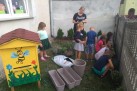 Na zdjęciu dzieci pod opieką osoby dorosłej Sadzą rośliny. Obok dzieci różne donice. Widać również żółty ul z rysunkiem kwiatów i pszczoły. 