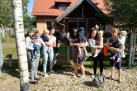 Kobiety trzymają dzieci na rękach. W zagrodzie znajduje się koń. W tle widać dom oraz drzewa.