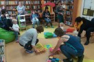Na zdjęciu widać grupę dorosłych i małych dzieci, prawdopodobnie w bibliotece, sądząc po regałach z książkami w tle. Dwie kobiety klęczą na podłodze, układając kolorowe, piankowe maty z puzzli. Wokół nich bawią się dzieci. Inni dorośli siedzą, obserwując lub trzymając na kolanach swoje dzieci.