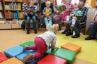 Zdjęcie pokazuje grupę dorosłych i małych dzieci biorących udział w spotkaniu w bibliotece lub podobnym miejscu wypełnionym książkami. Kilka osób dorosłych siedzi z dziećmi na kolanach, czytając m.in. im książki. W tle widoczny jest regał z licznymi książkami. Na pierwszym planie mała dziewczynka w różowych spodniach bawi się na kolorowych poduszkach.