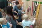 Na zdjęciu widać grupę dorosłych i małych dzieci, którzy bawią się przy interaktywnej tablicy sensorycznej. Dzieci są aktywnie zaangażowane w eksplorowanie różnych elementów na tablicy, takich jak kolorowe sznurki z koralikami i pomponami oraz sznurowanie buta. Dorośli nadzorują i pomagają dzieciom w zabawie.