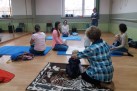 Zdjęcie przedstawia zajęcia dla rodziców z małymi dziećmi, prawdopodobnie w sali do ćwiczeń. Na zdjęciu widać kilka kobiet z dziećmi, siedzących na matach na podłodze. Jedna z kobiet, stojąca w głębi sali, wydaje się prowadzić zajęcia. Uczestnicy są w swobodnych pozycjach, co sugeruje, że mogą to być zajęcia ruchowe.