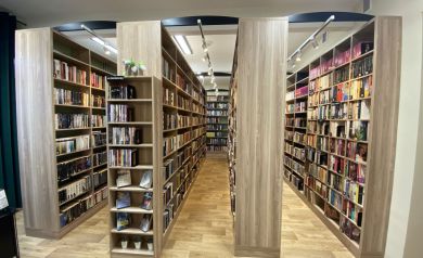 Na zdjęciu widać wnętrze nowoczesnej biblioteki lub księgarni. Pomieszczenie jest jasne, z drewnianą podłogą i równymi rzędami wysokich regałów w kolorze jasnego drewna. Półki są wypełnione książkami ułożonymi w porządku, a nad alejkami znajdują się nowoczesne lampy sufitowe zapewniające dobre oświetlenie. Na jednym z regałów stoi mała dekoracja w postaci pluszowej rośliny.