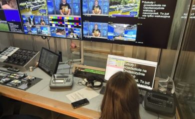 Na zdjęciu pomieszczenie z wieloma ekranami znajdujące się w budynku TVP 3 Opole. Przy komputerze siedzi młoda dziewczyna w fioletowej koszulce projektowej. - powiększ