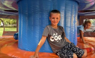 Na zdjęciu chłopiec siedzi na dmuchanej, kolorowej atrakcji – w środku dużego dmuchańca. Ma na sobie koszulkę z napisem „MR COOL” i uśmiecha się do aparatu. W tle widać drugie dziecko, które również się bawi.