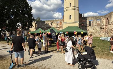 Zdjęcie przedstawia jedną z atrakcji wydarzenia „Zakręcona Wakacyjna Meta”, odbywającego się na tle ruin zabytkowego zamku z wieżą. W centrum znajdują się zielone namioty ozdobione kolorowymi balonami, przy których zebrała się spora grupa uczestników – głównie rodziny z dziećmi. Pogoda dopisuje – niebo jest niemal bezchmurne.