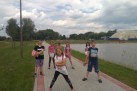 Na zdjęciu widać grupę dzieci, które pozują do zdjęcia na ścieżce spacerowej. Na pierwszym planie znajduje się siedmioro dzieci w różnym wieku. Jeden chłopiec trzyma w rękach czerwoną piłkę. Dzieci stoją na chodniku wyłożonym kostką brukową, który biegnie wzdłuż brzegu zbiornika wodnego. Po drugiej stronie ścieżki znajduje się trawiasty teren z drzewami. W oddali widać budynek i fragmenty infrastruktury rekreacyjnej. Niebo jest częściowo zachmurzone, ale światło wskazuje na pogodny dzień.