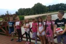 Na zdjęciu siedmioro dzieci. Jeden z chłopców trzyma piłkę biało-czerwoną. Dzieci stoją na drewnianym pomoście przy zbiorniku wodnym. W oddali widać prawdopodobnie park.