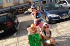 Na zdjęciu trzy dziewczynki w dużych kapeluszach i okularach przeciwsłonecznych pozują na słonecznym placu, trzymając kolorowo ozdobione torby. W tle widać samochody i zabudowę miejską.