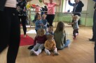 Na fotografii widać grupę małych dzieci oraz dorosłych w sali tanecznej z lustrami. Na pierwszym planie, na drewnianej podłodze, siedzi kilkoro małych dzieci. W tle dorośli (głównie kobiety) trzymają dużą, kolorową, tęczową chustę animacyjną (materiał) nad głowami innych, stojących dzieci, które aktywnie uczestniczą w zabawie. Otoczenie obejmuje dużą, jasną salę z lustrami na ścianie w tle, grzejnikiem oraz kocami na podłodze.