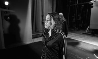 Fotografia czarno-biała przedstawia kobietę, stojącą na scenie. Kobieta o kręconych włosach, ubrana w czarną bluzę dresową z białymi paskami na rękawach, patrzy w lewą stronę z intensywnym wyrazem twarzy. Za nią, w tle, znajduje się druga, niewyraźna postać. Otoczenie sugeruje teatralną scenę, z ciemnymi zasłonami, elementami oświetlenia scenicznego i pustą przestrzenią.
