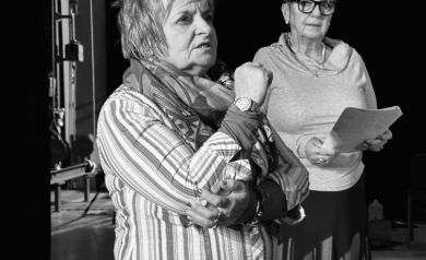 Zdjęcie przedstawia dwie starsze kobiety, prawdopodobnie podczas próby teatralnej. Kobieta po lewej ma krótkie, rozczochrane włosy i intensywny wyraz twarzy. Ubrana jest w koszulę w paski, szalik i zegarek na prawym nadgarstku. Jej prawa pięść jest zaciśnięta, co sugeruje emocjonalne zaangażowanie lub energiczną gestykulację. Kobieta po prawej stoi obok, ubrana w szary sweter i długą, ciemną spódnicę. Nosi okulary w prostokątnych oprawkach i trzyma w rękach plik papierów, prawdopodobnie scenariusz. Jej postawa jest bardziej stonowana w porównaniu do drugiej kobiety. Fotografia jest czarno-biała.