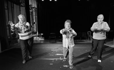 Na zdjęciu w czerni i bieli widać trzy starsze kobiety na scenie teatralnej. Kobiety wydają się być w trakcie próby teatralnej, przyjmując dynamiczne pozy z wyciągniętymi rękami. Akcja rozgrywa się na scenie z ciemnym tłem, czarnymi zasłonami i widocznym oświetleniem scenicznym u góry. Podłoga sceny jest ciemna z widocznymi liniami taśmy klejącej. W tle widoczne są puste krzesła oraz elementy konstrukcji scenicznej.
